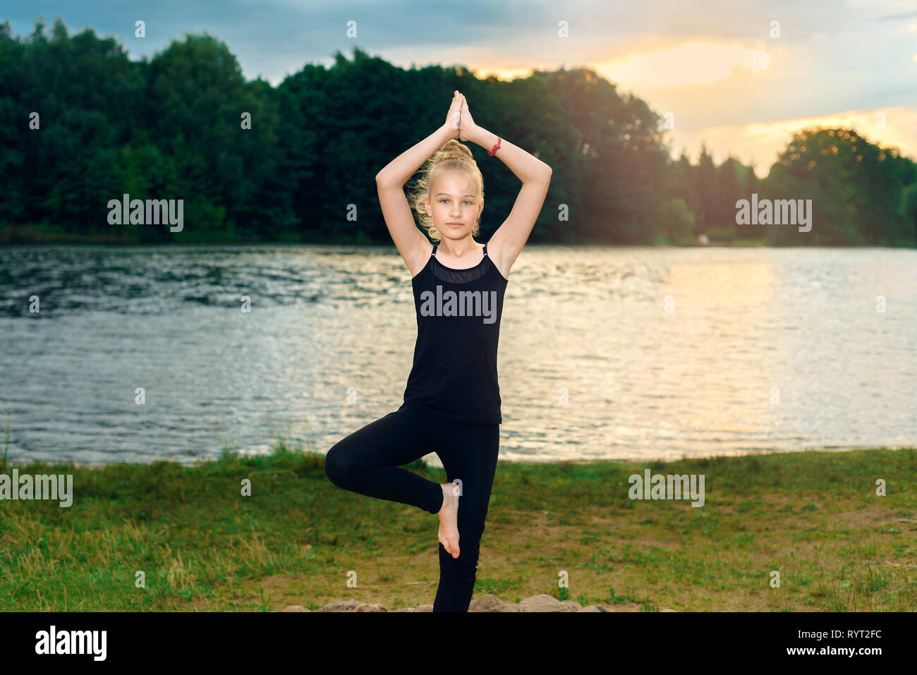 young blonde girl in black T-shirt and leggings in lotus position, namaste doing yoga, pilates by the lake at sunset. concept of healthy life, inspira Stock Photo