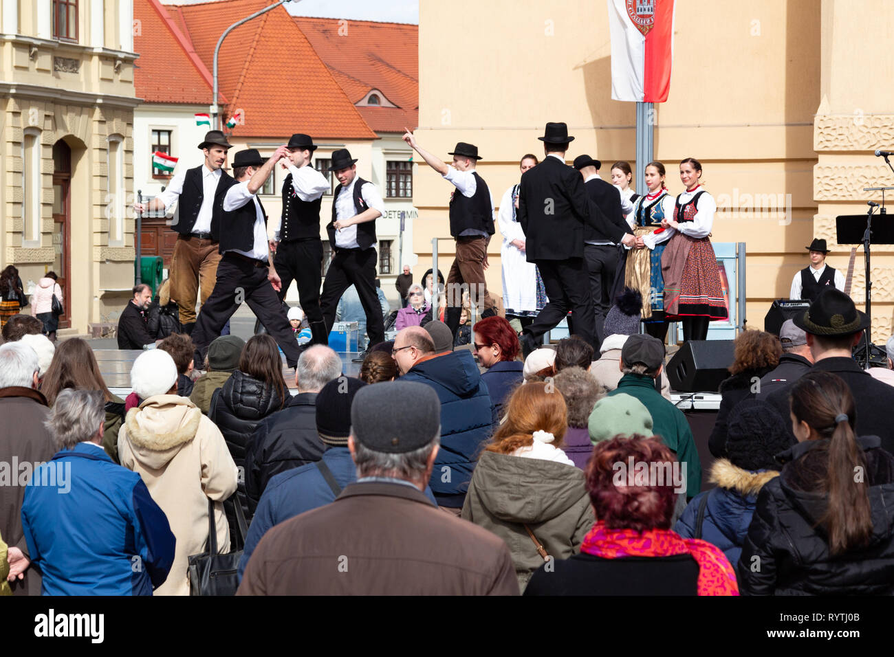 Sopron, Hungary. 15th Mar, 2019. Group of male folk dancers performs traditional Hungarian dances in circle on stage at Petőfi Square, Sopron, Hungary. Girls of the folk dance group cling together at the edge of the stage watching. Credit: Wahavi/Alamy Live News Stock Photo