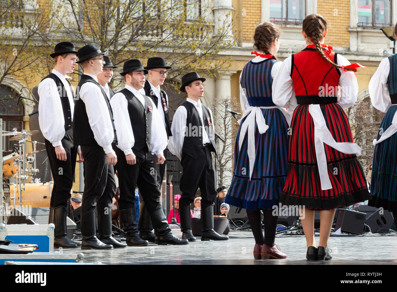 Sopron, Hungary. 15th Mar, 2019. A group of folk dancer lads stand and sing on stage at Petőfi Square, Sopron, Hungary. Girls in traditional costume stand on the right. Credit: Wahavi/Alamy Live News Stock Photo