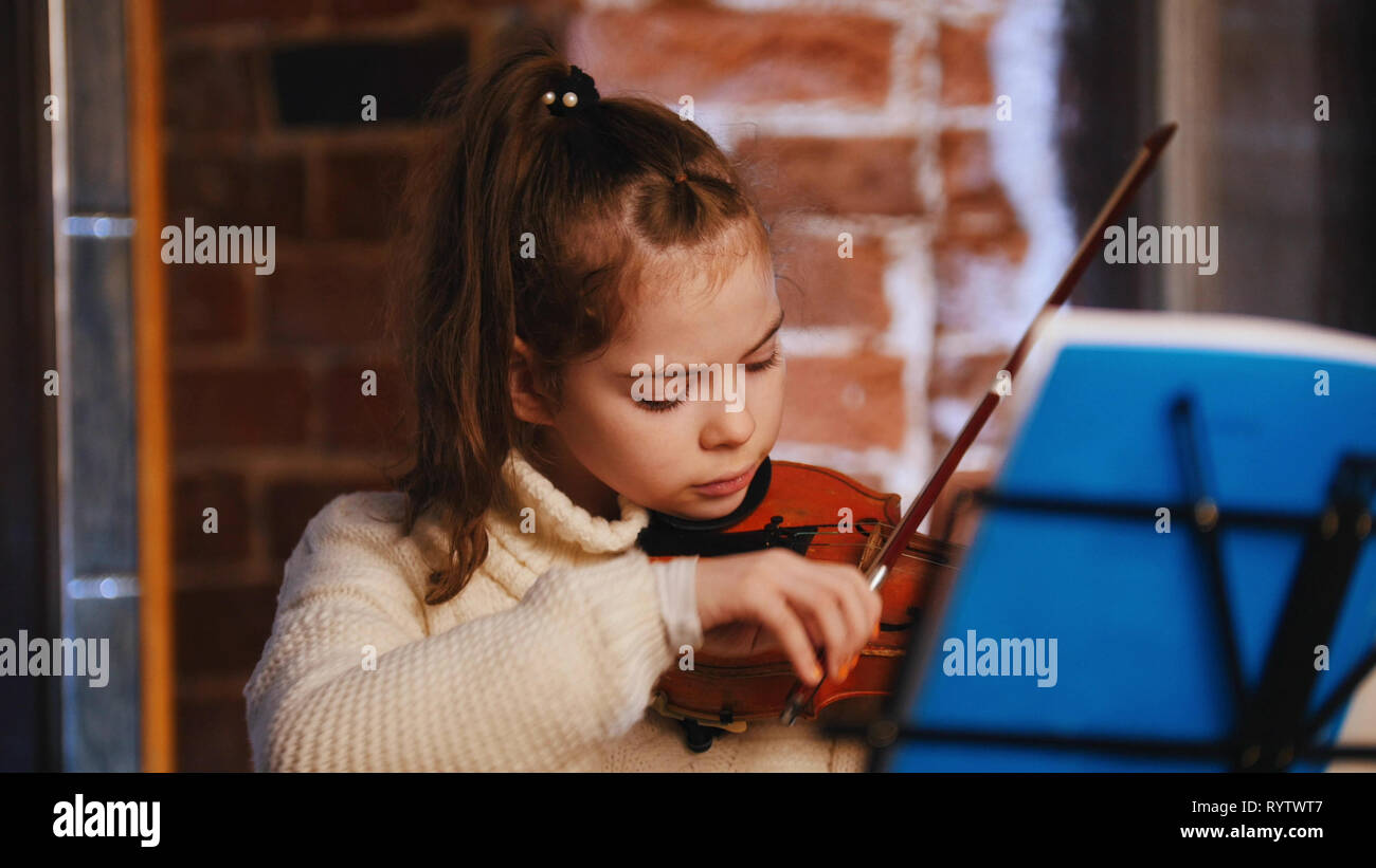 A little concentrated girl in white sweater playing violin by notes ...