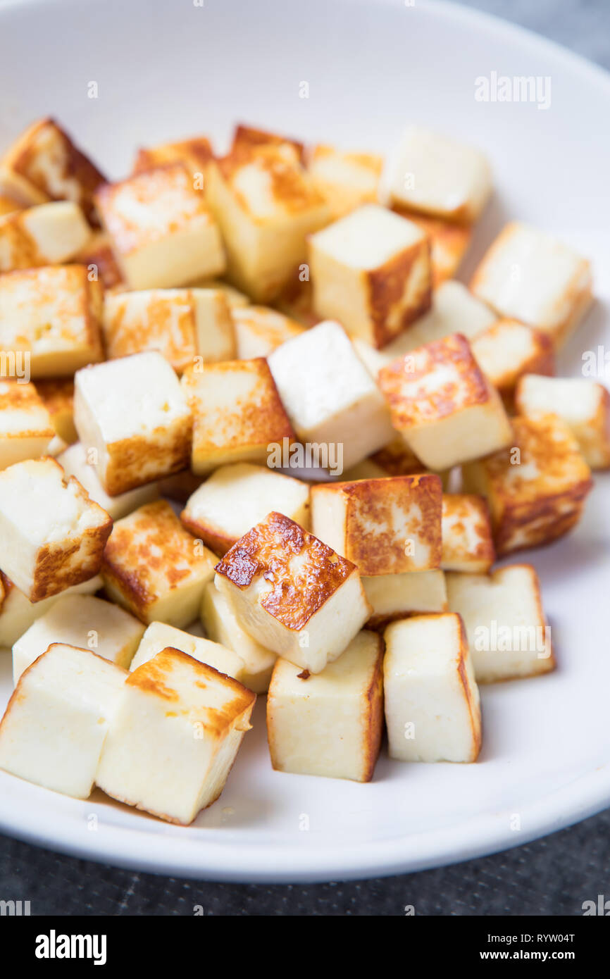 Fried small cubes of paneer in the foreground on a white plate. Close up picture of small fried cubes of cottage cheese. Pan-fried paneer cubes. Stock Photo