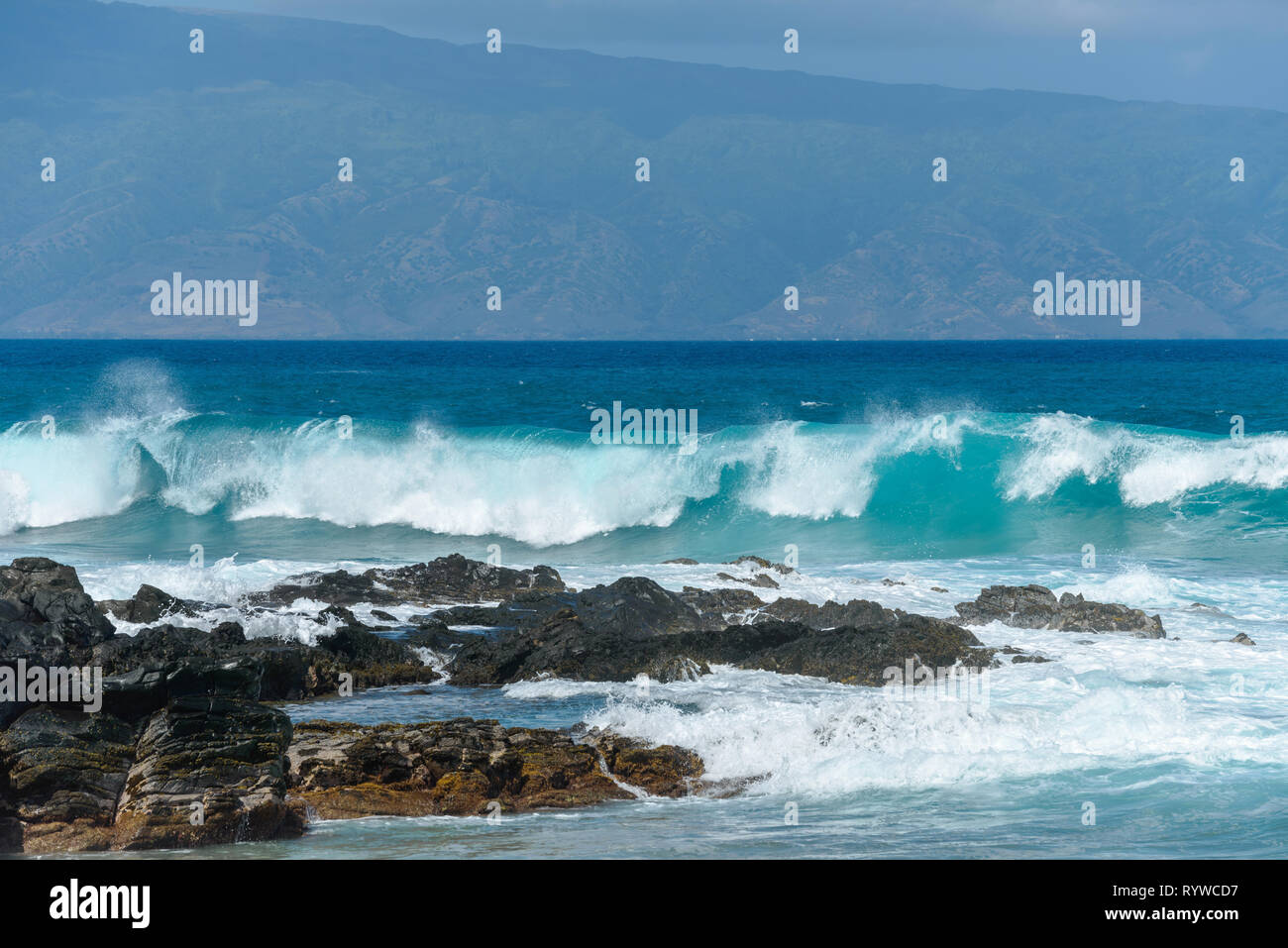 Breaking Waves - Big breaking blue waves rushing towards black rocky coast, with a tropical island in the background. Maui, Hawaii, USA. Stock Photo