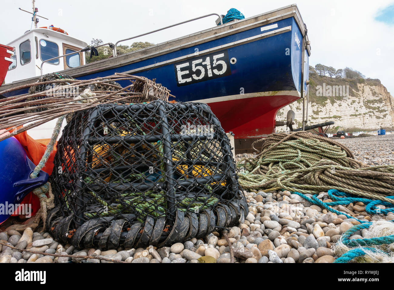 A fishing boat and lobster pot sit on the shingle beach in Beer, Devon, England. Ropes, a fender and a lobster pot are sitting near the fishing  boat. Stock Photo