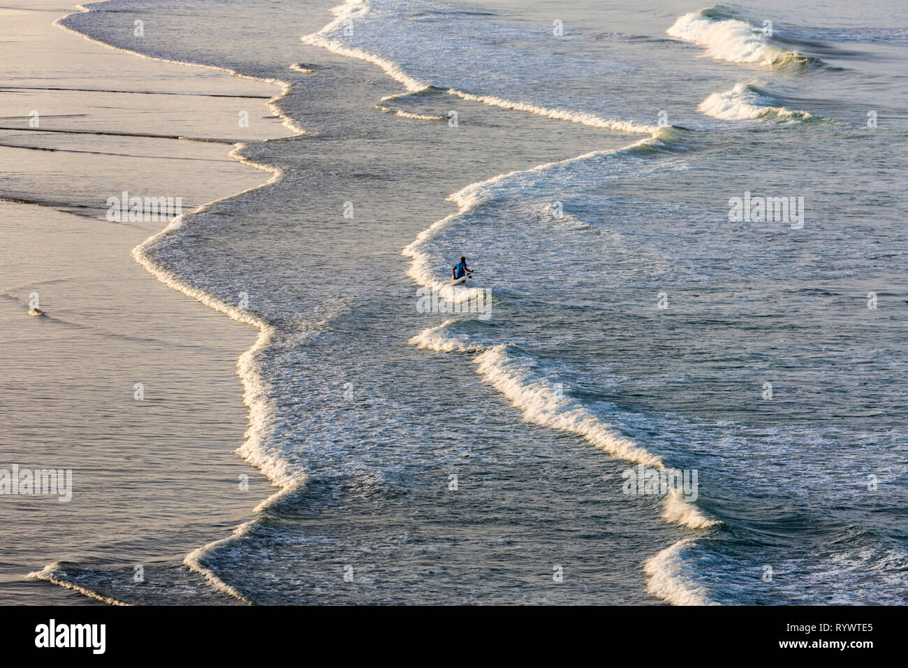 slow incoming Pacific waves at sunset with reflecting sunlight and a lonley surfer, Northland, New Zealand Stock Photo