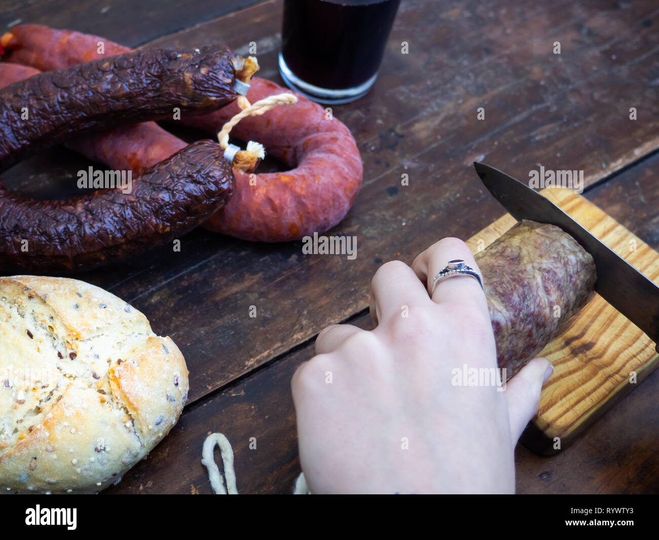 One person cutting an Iberian pork sausage on an old wooden board with an antique knife and rustic bread and a glass of red spanish wine Stock Photo