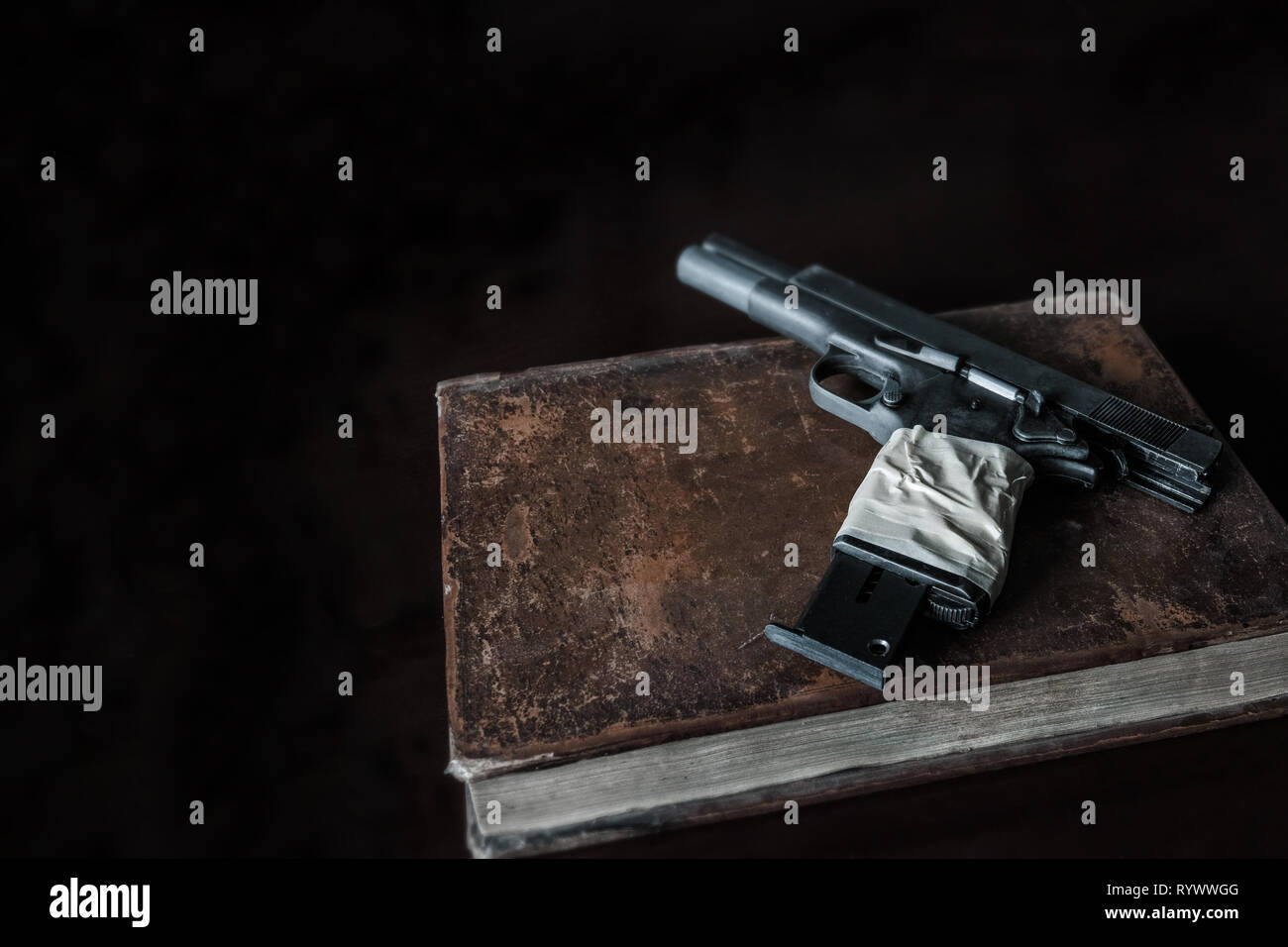 On an old book, probably a book of wisdom, a pistol whose handle is wrapped with tape. Low key, copy space. Stock Photo
