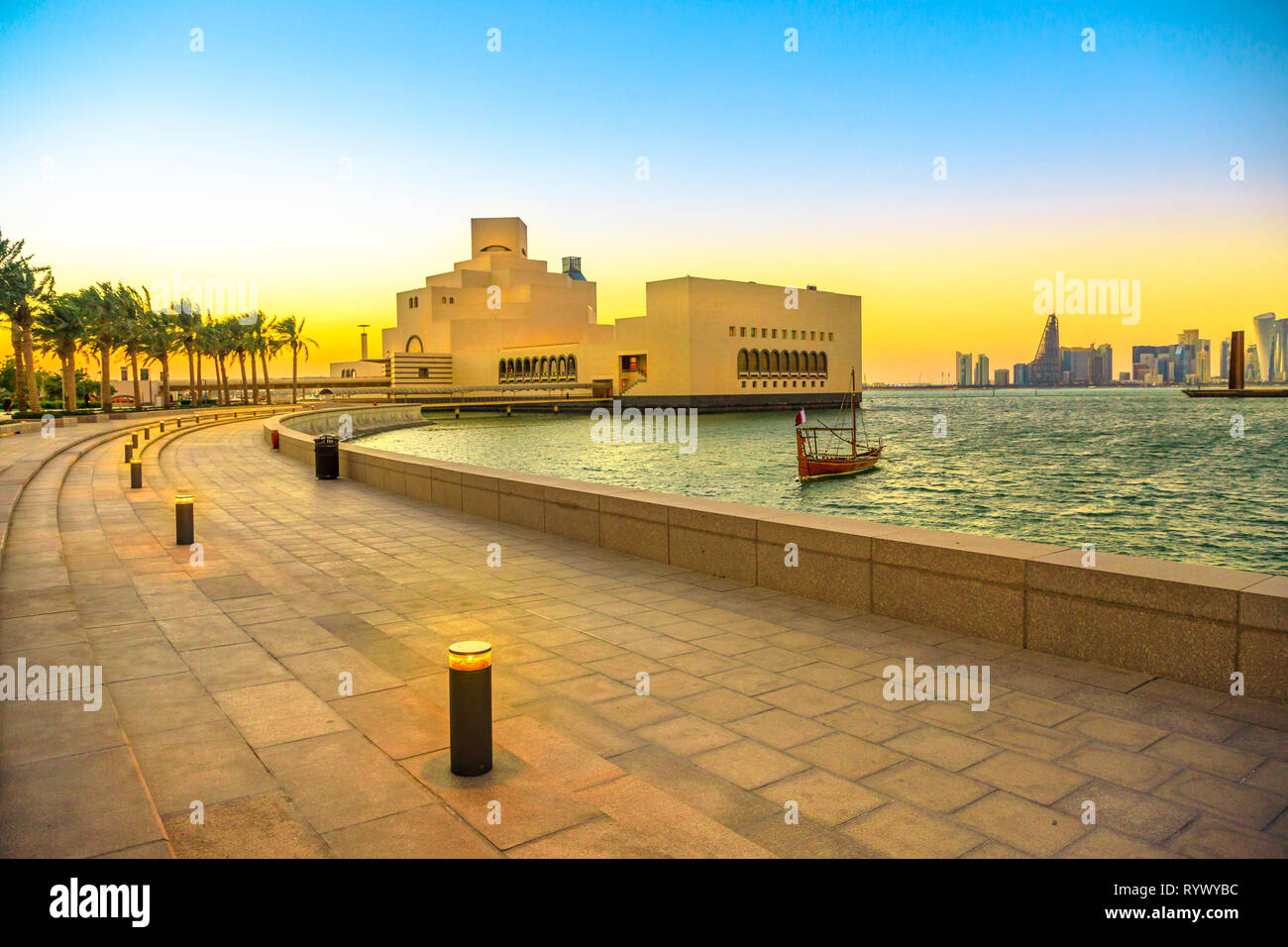 Seafront walkway with palm trees along Doha Bay with dhow and skyscrapers of Doha West Bay skyline at sunset. Urban cityscape. Qatari capital in Stock Photo