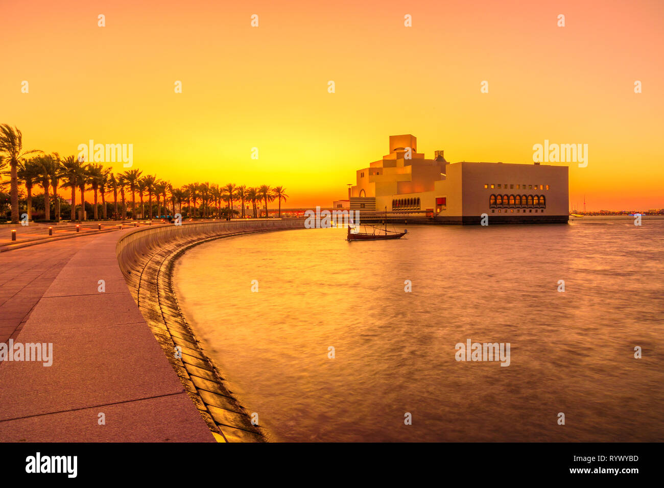 The walkway with palm trees along Doha Bay. Museum and dhow at sunset