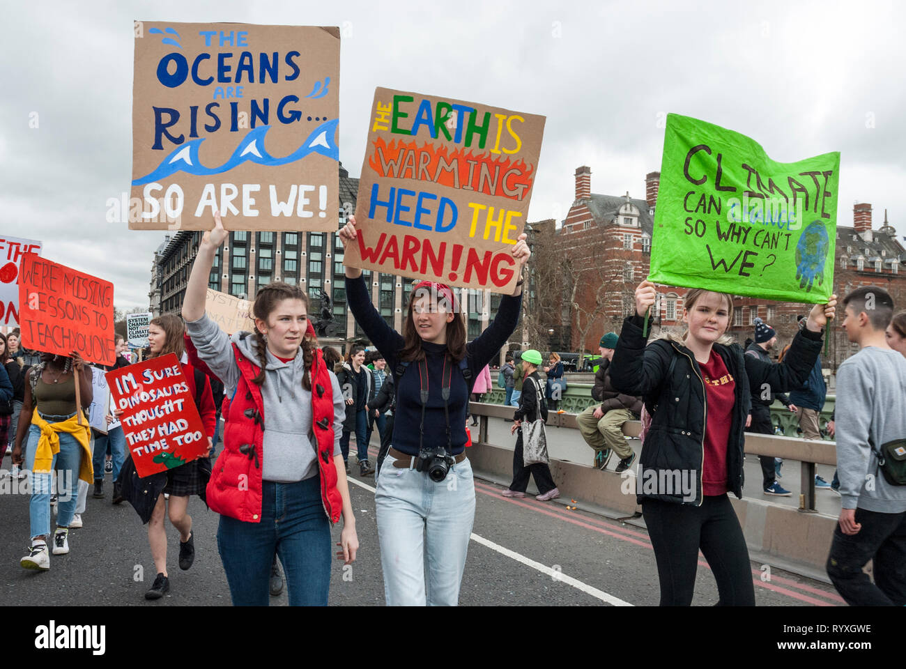 London, UK. 15th March, 2019. School students campaigning against climate change protest outside Parliament. Credit: Maggie sully/Alamy Live News. School children on strike to protest at climate change march over Westminster bridge, outside Parliament, London. In the foreground three young women students carry colourful homemade banners 'The oceans are rising...so are we!', 'The earth is warming, heed the warning' and 'Climate can change so why can't we?'. Part of the worldwide 'FridaysforFuture' protest. Stock Photo