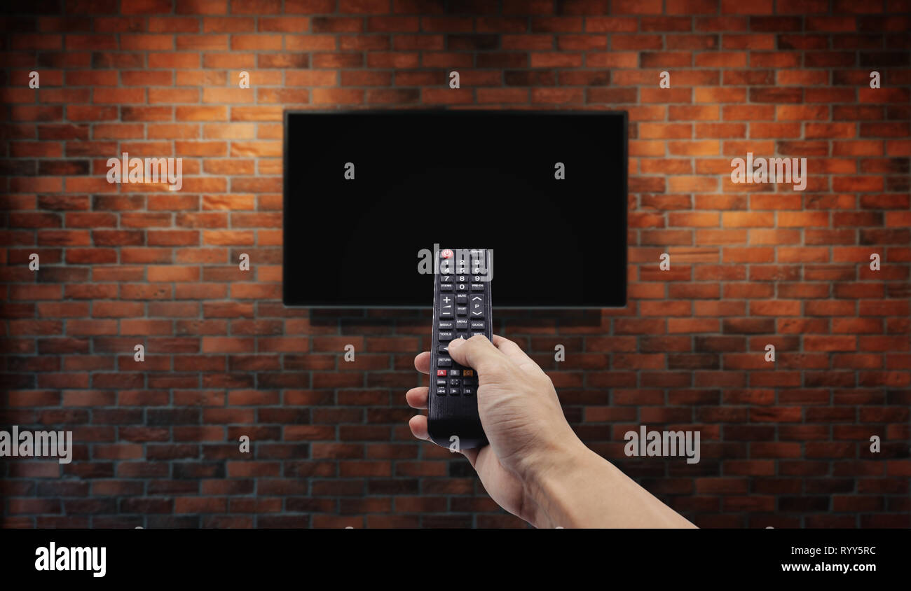 Television on brick wall with hand using remote control Stock Photo