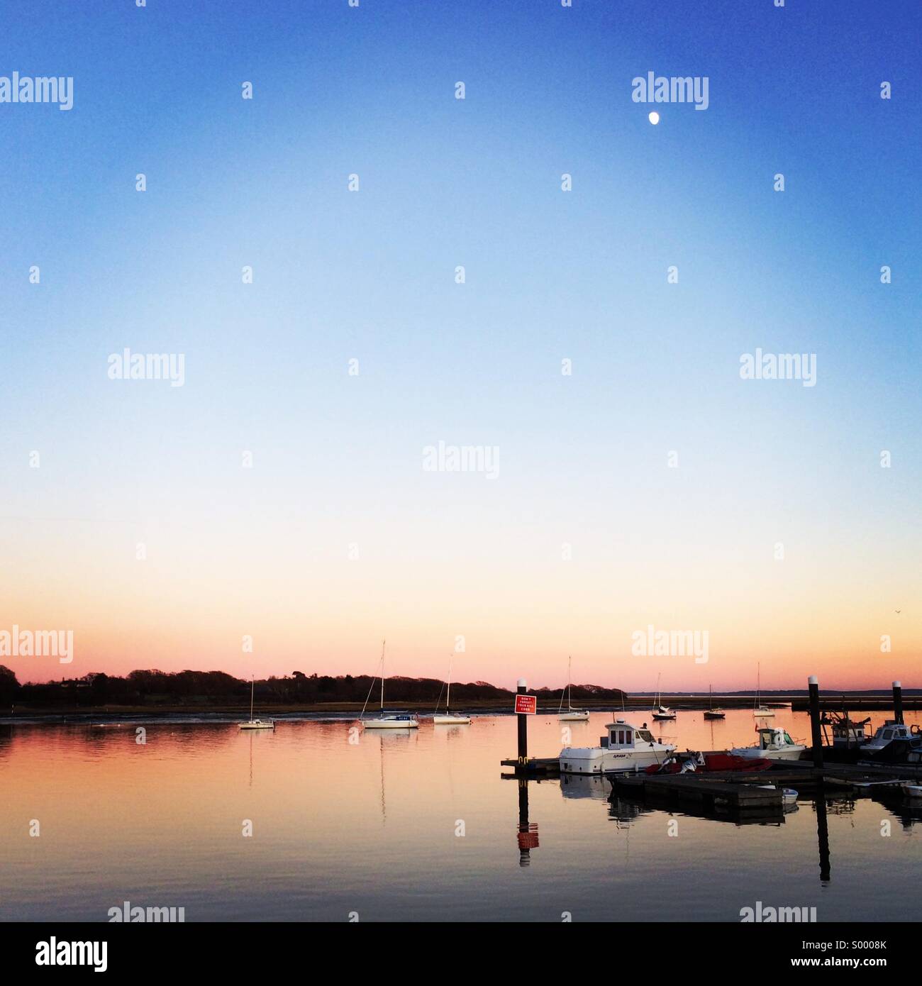 Sunset at the Royal Yacht Club at Lymington.  The moon rises as the sun sets and the water turns to glass. Sailboats moor in the shelter of the harbour. - Smartphone Captured Stock Image