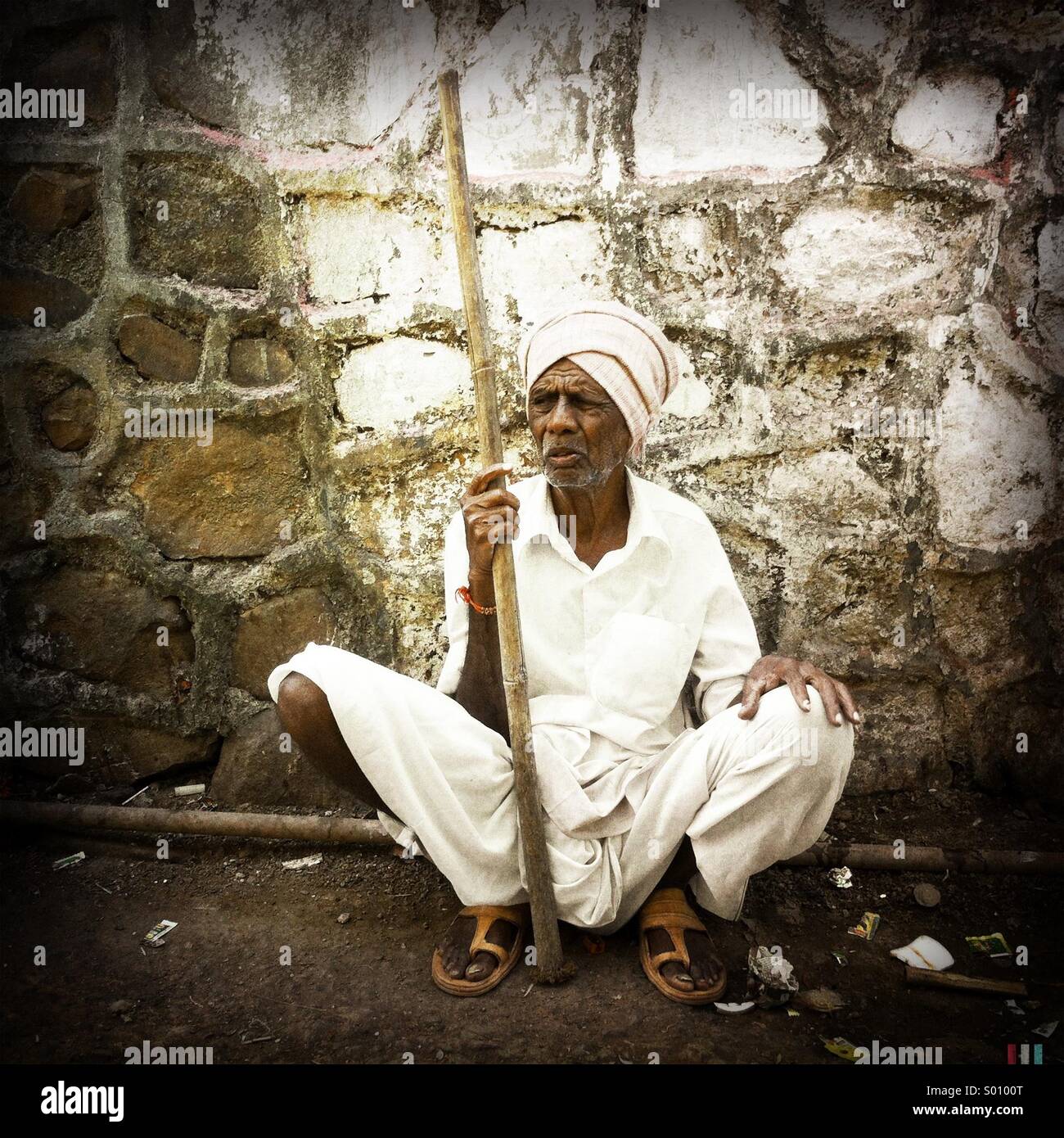An indian man sitting on street. Man is holding a wooden stick. - Smartphone Captured Stock Image