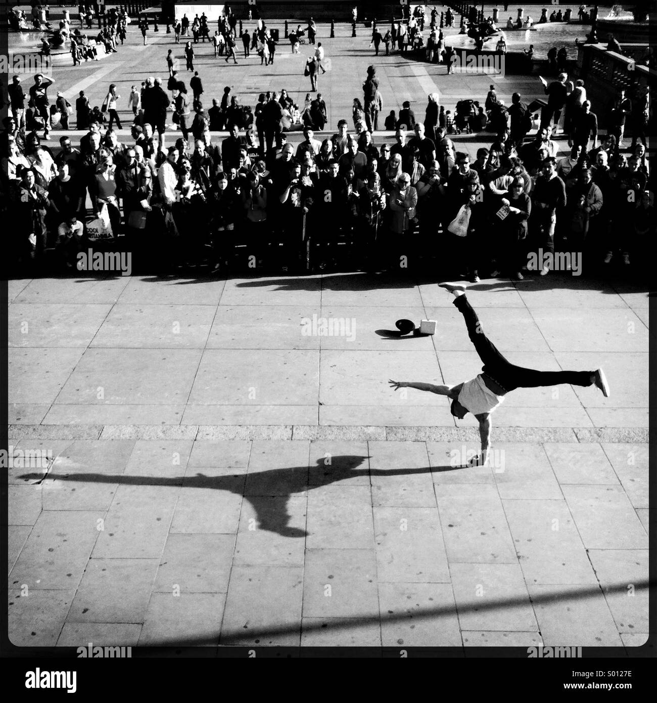 Black and white image of a street performer doing a handstand in front of a crowd of onlookers as the late afternoon sun casts a shadow. Stock Photo