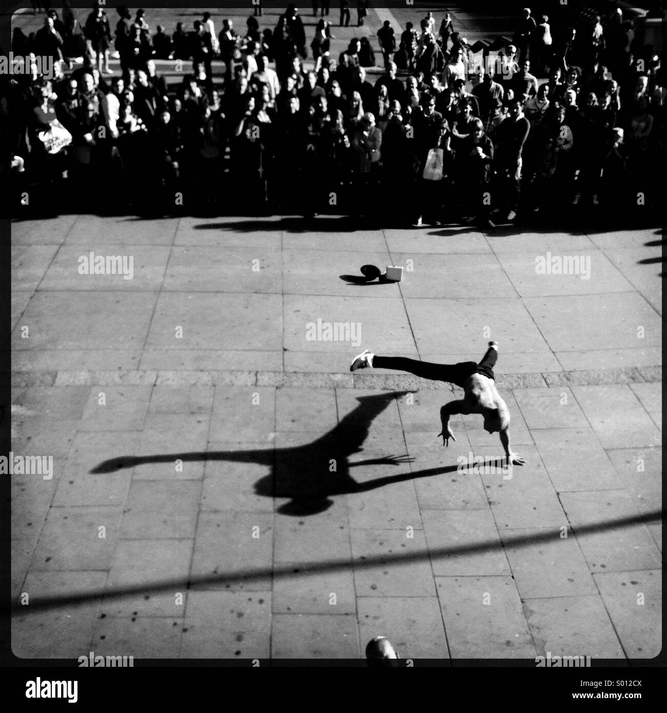 Black and white image of a street performer doing a handstand in front of a crowd of onlookers as the late afternoon sun casts a shadow. Stock Photo