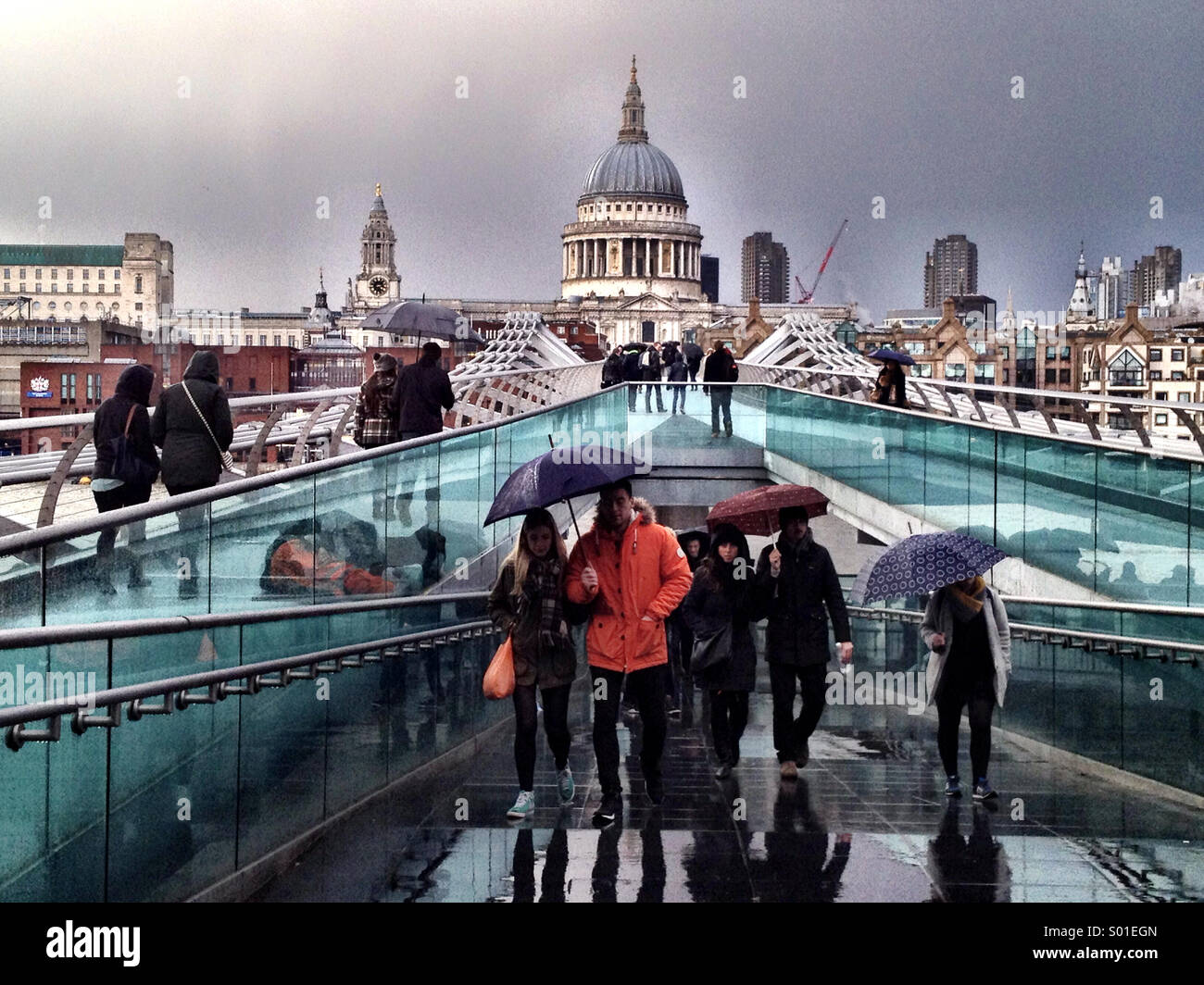 People carrying umbrellas cross the Millenium Bridge in London on a wet spring day - Smartphone Captured Stock Image