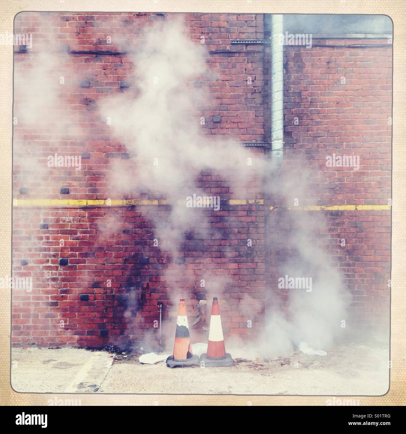 Steam or smoke rises from a hole in the ground surrounded by traffic cones. There is a red brick wall as a backdrop Stock Photo