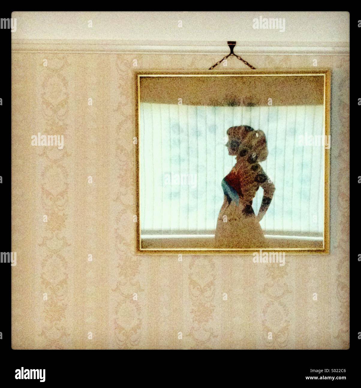 Reflection of the silhouette of a woman in the glass of a framed artwork hanging from a picture rail on a wall with patterned wallpaper-a self portrait. - Smartphone Captured Stock Image