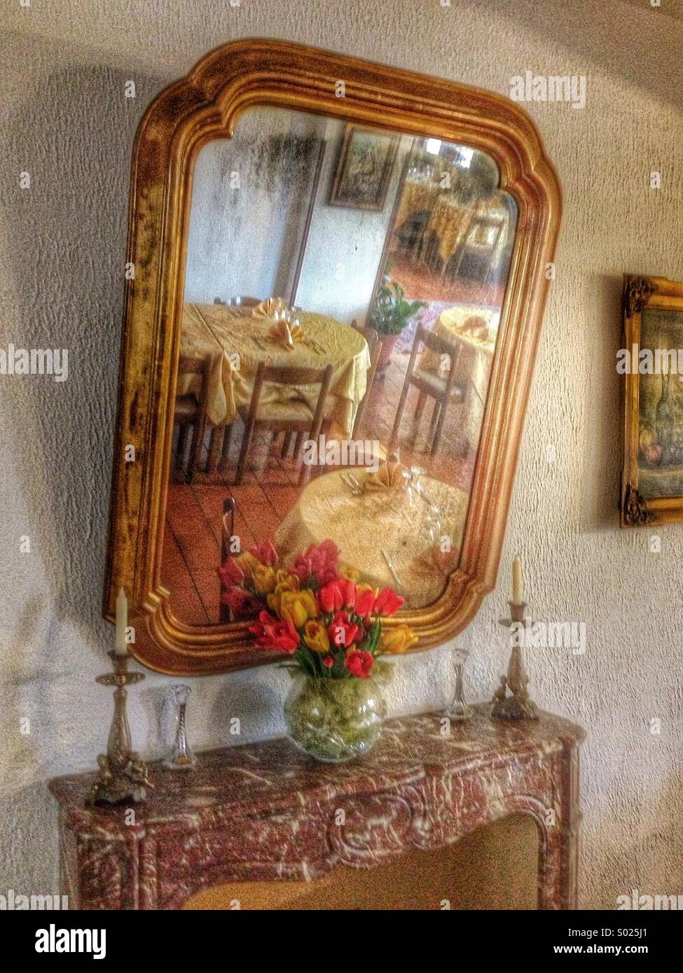 Reflections of restaurant tables and chairs in a mirror, above a mantelpiece. Gold edged mirror frame, white wall, tulips in a vase and candles either side of the mantelpiece. Grainy effect. - Smartphone Captured Stock Image