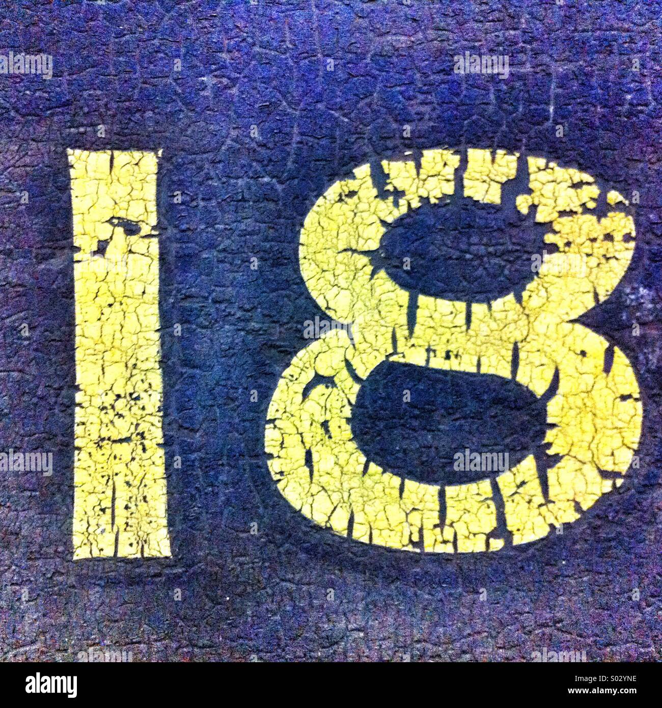 Bright yellow number 18 painted on a cracked blue surface in a public space during daylight - Smartphone Captured Stock Image