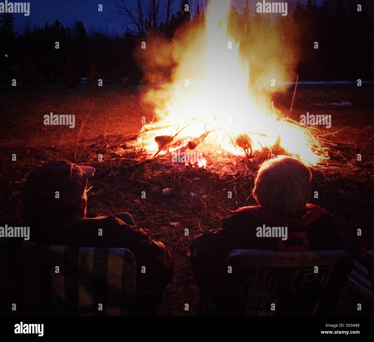 Watching The Bonfire, Two Mature Men Sat On Deck Chairs Enjoying A Bonfire Stock Photo