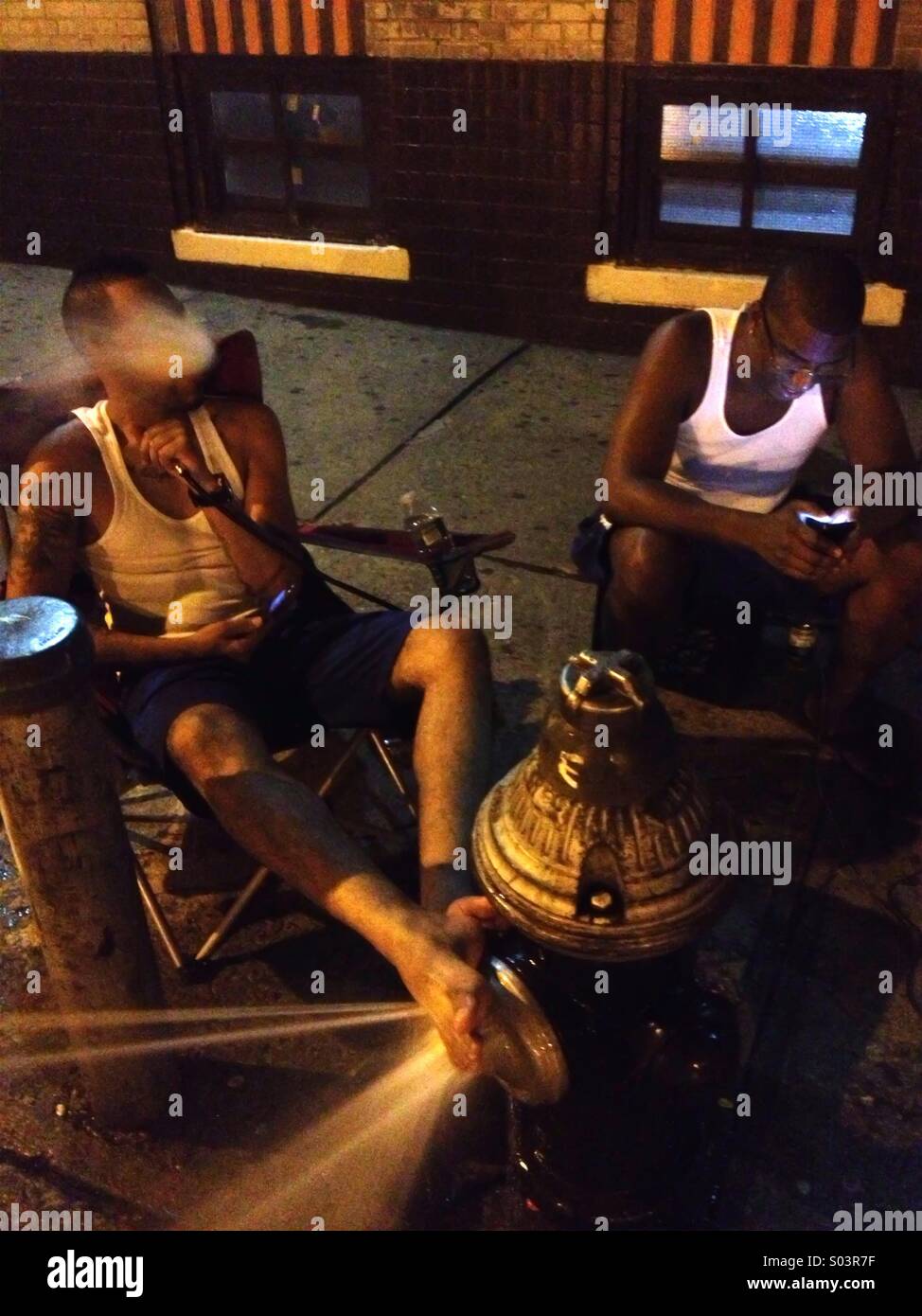 Two men hanging out on street on hot summer night, smoking hookah pipe, cooling feet off on fire hydrant - Smartphone Captured Stock Image