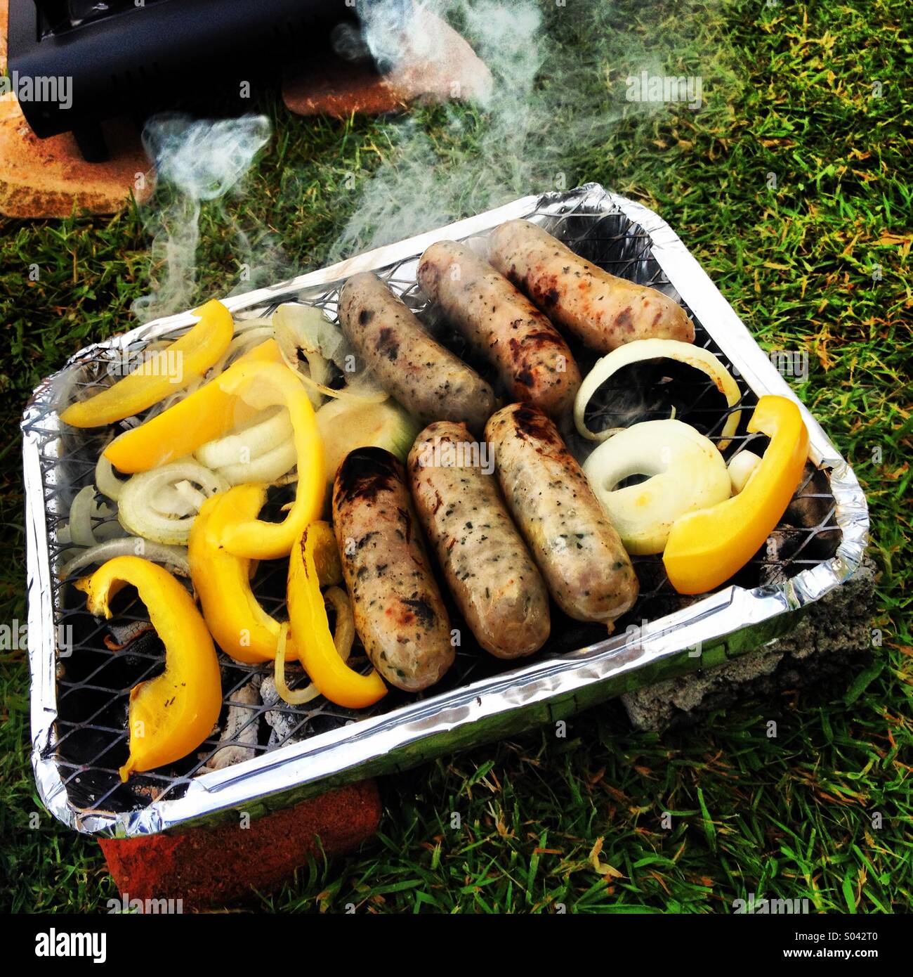 Sausages, onions and peppers in disposable barbecque, while camping. Stock Photo