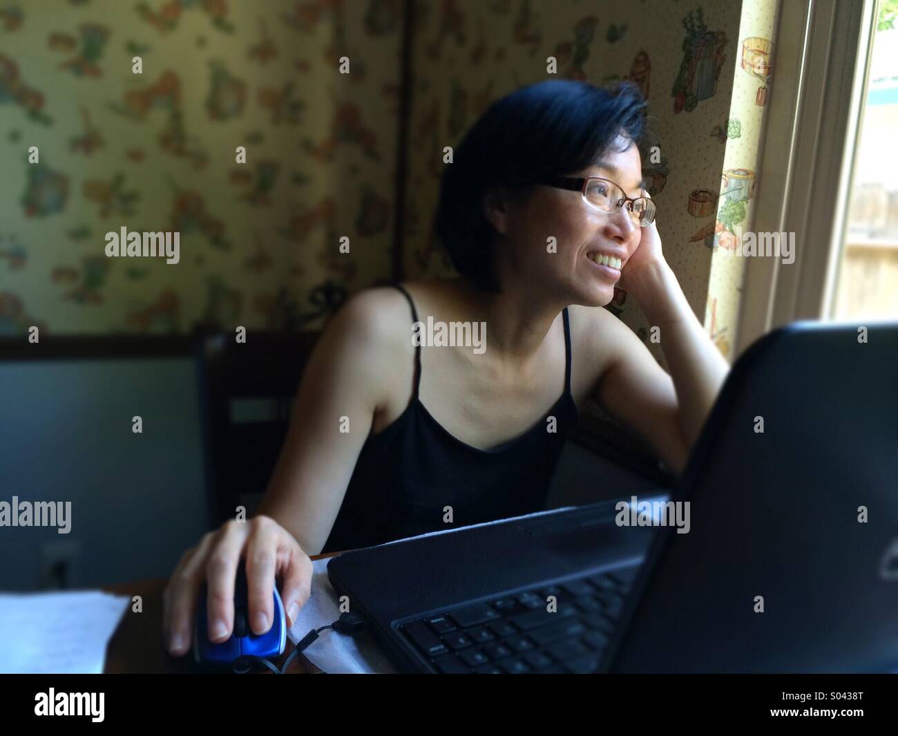 Asian woman smiling and looking out a window while using a laptop computer at home Stock Photo
