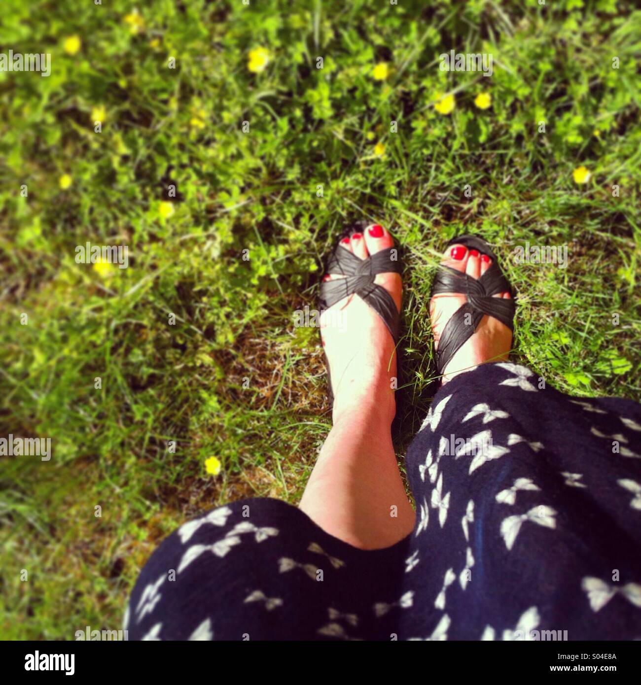 Looking down at feet in sandals and a summer dress standing on a LAN with small yellow flowers Stock Photo