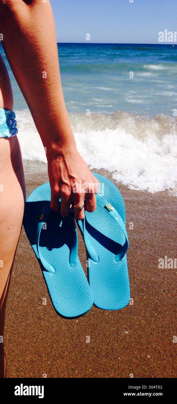 Woman on beach holding blue flip flops, sea in background - Smartphone Captured Stock Image