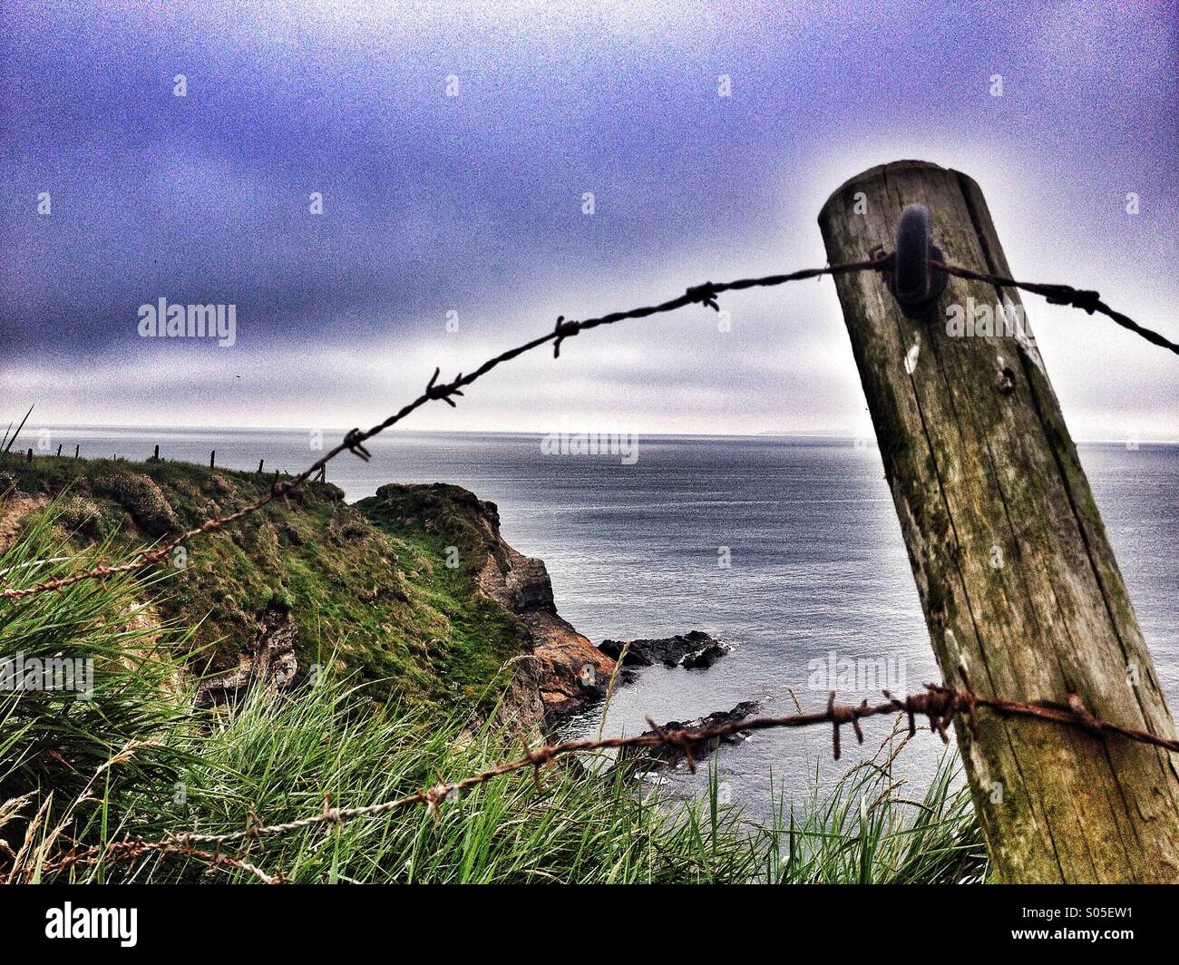 Broken barbed wire fence with cliff top cove in background Stock Photo