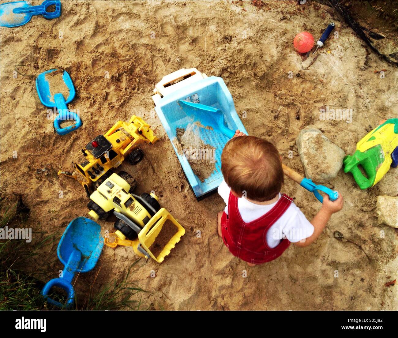 Toddler boy plays with a shovel and trucks in a large sandbox. Stock Photo