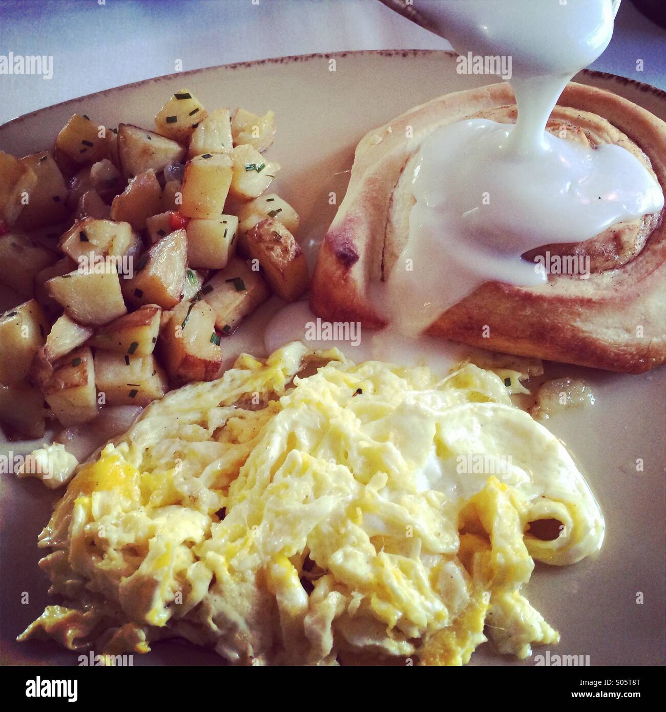 A hearty breakfast of scrambled eggs. Potatoes and a cinnamon roll over which thick white icing is being poured - Smartphone Captured Stock Image