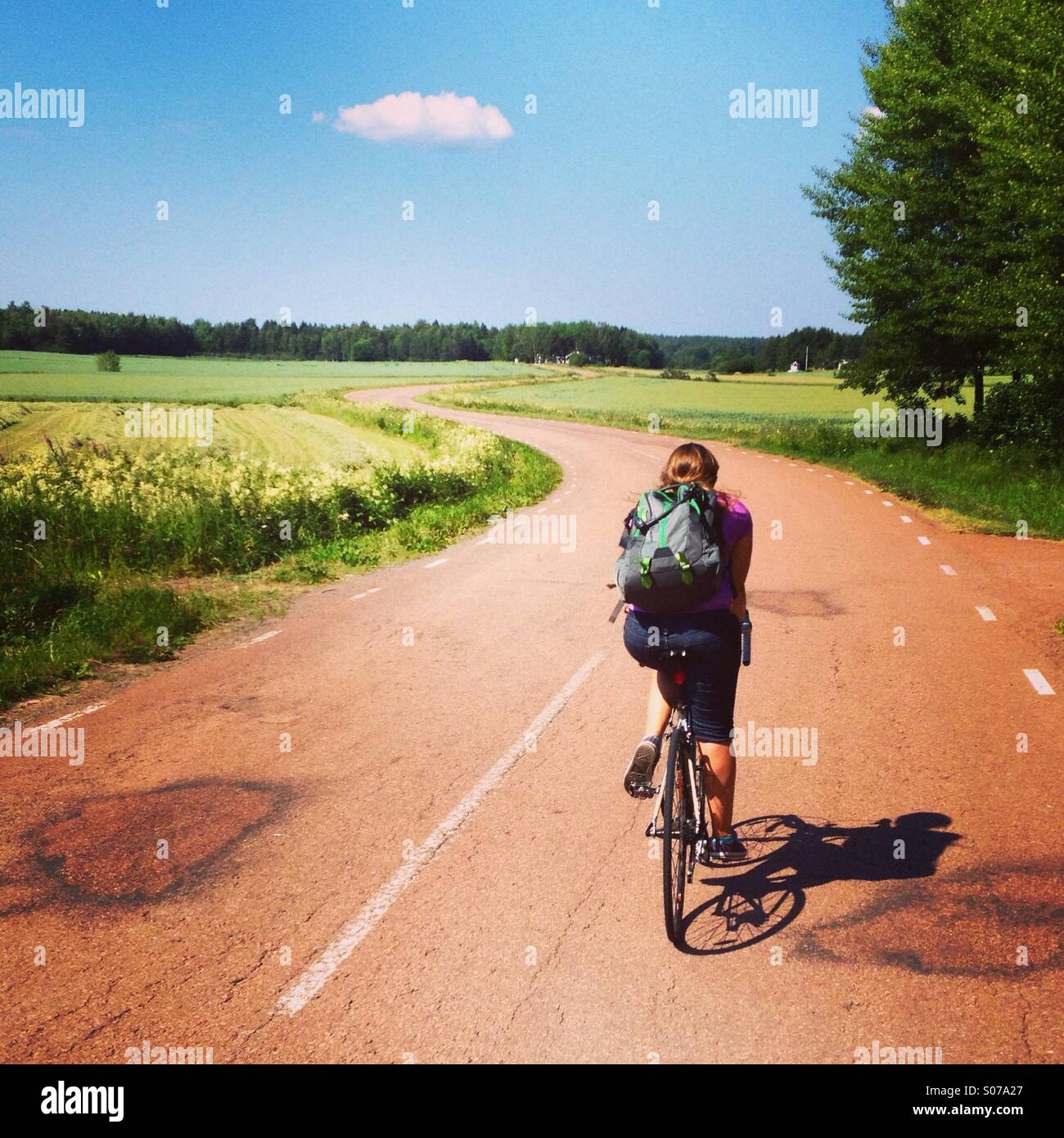 A woman on a cycling holiday on a snaking traditional red road in the Nordic Finnish archipelago of Åland - Smartphone Captured Stock Image