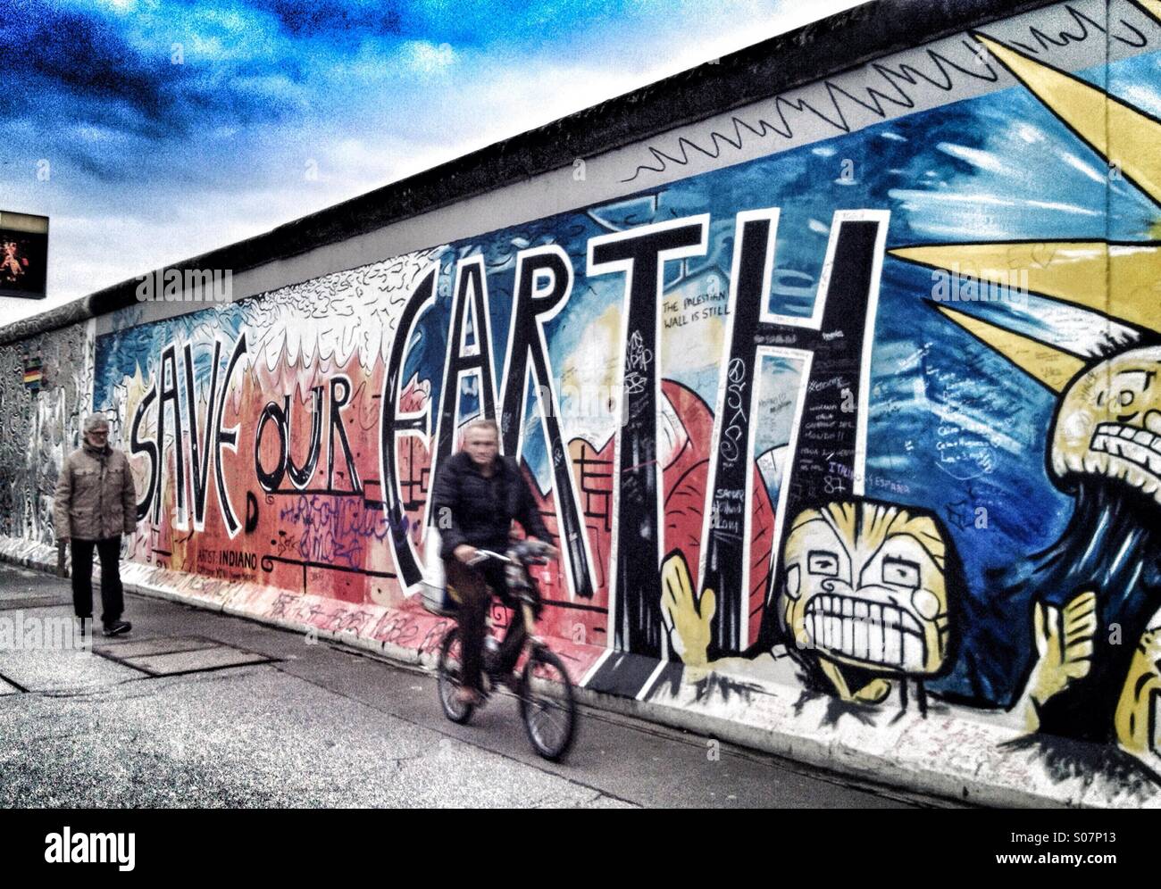 'Save our Earth'. A man cycles past a remnant of the Berlin Wall which has been painted with an environmental message - Smartphone Captured Stock Image