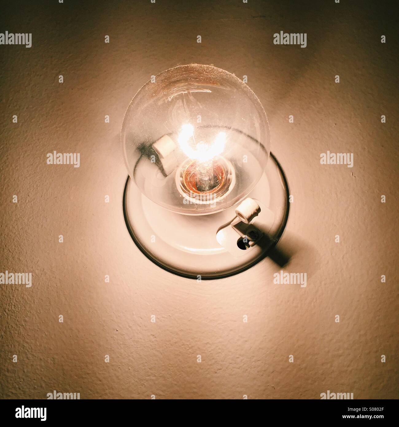 A single, large, clear, illuminated tungsten lightbulb mounted on a wall. Stock Photo