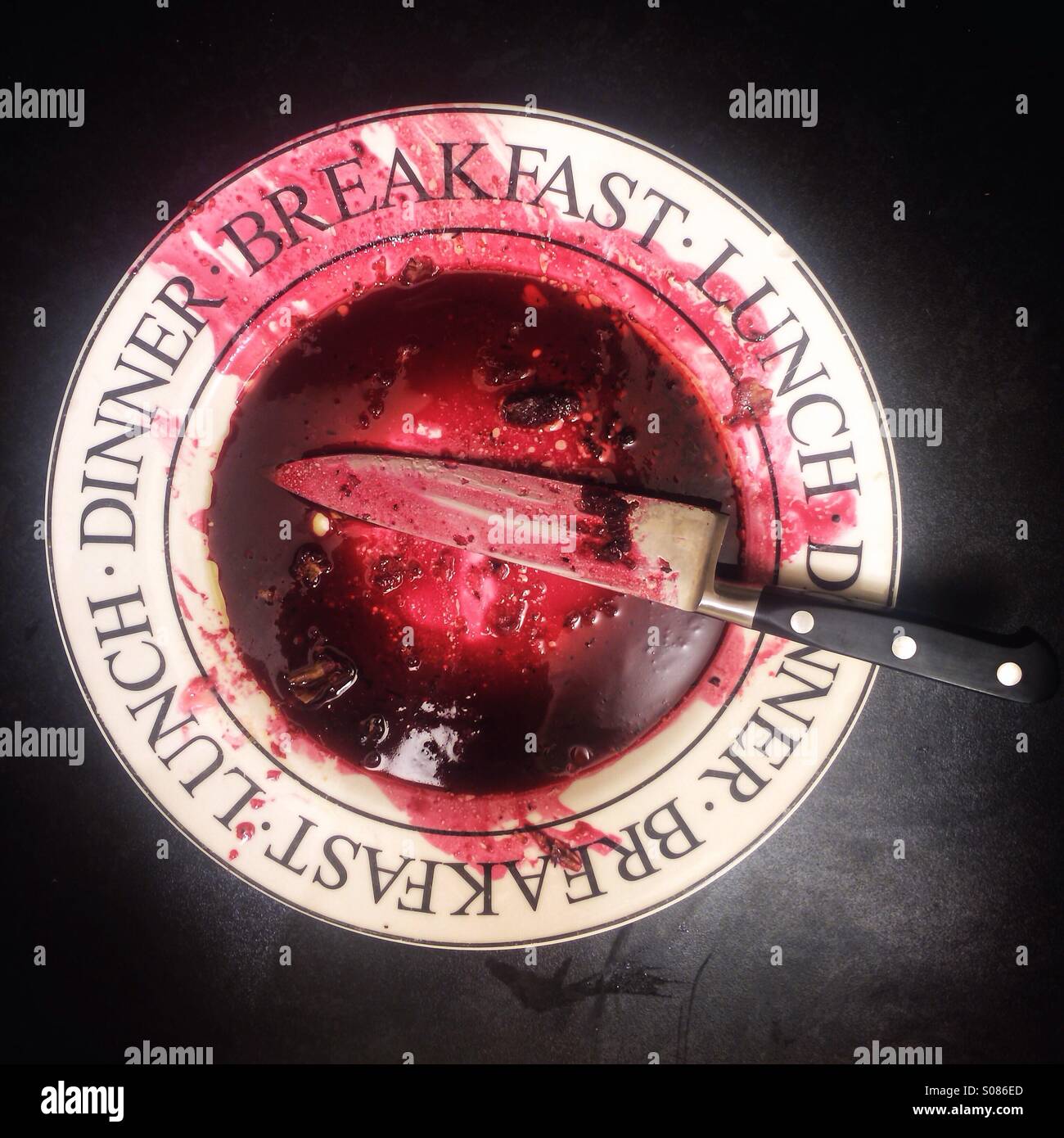 A white plate with black lettering stained with beetroot juice which looks like blood. On the plate is a sharp knife. Stock Photo