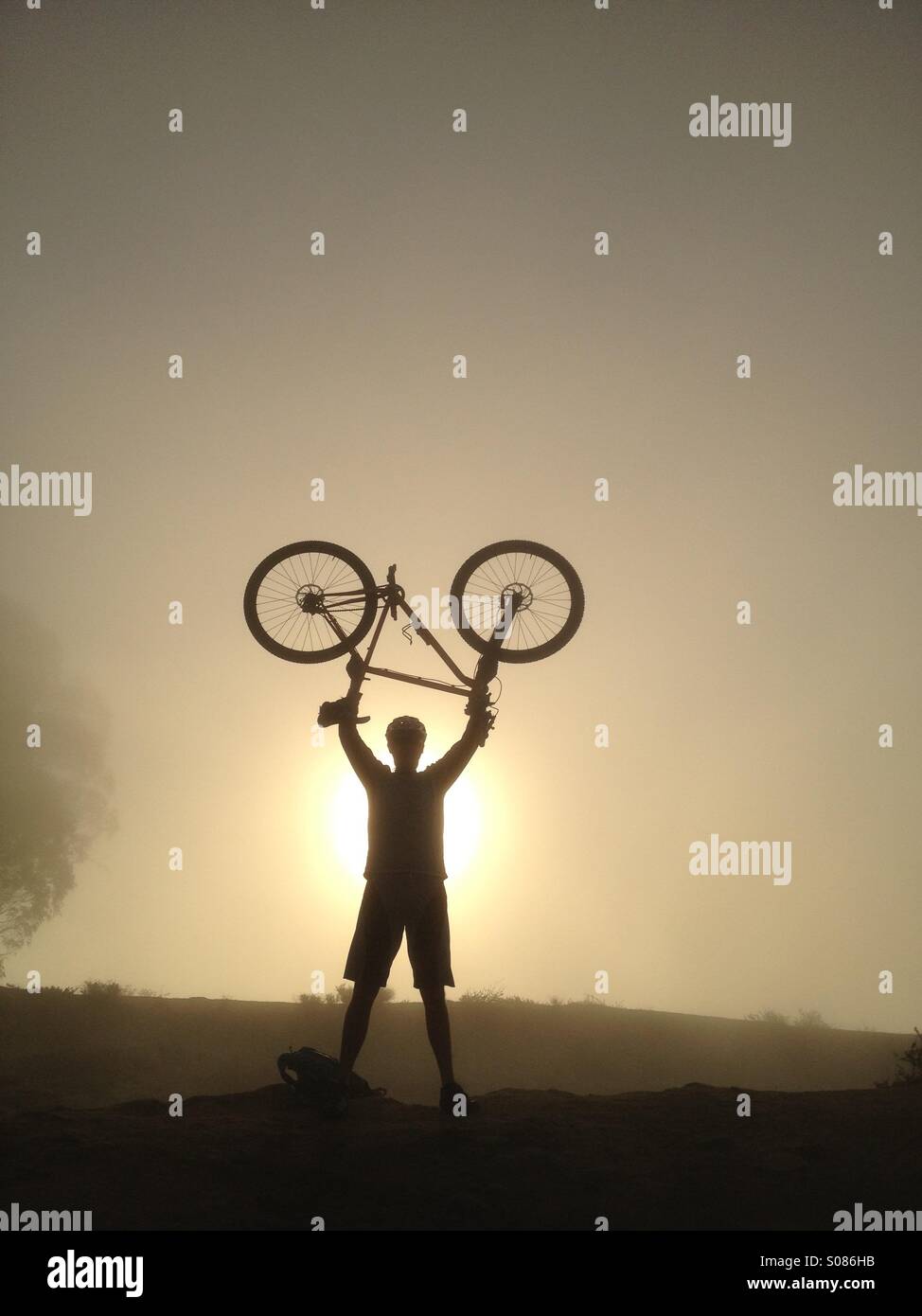 Silhouette in the sun a Mountain biker holds his bike over his head during a foggy misty morning ride in Laguna beach California. Stock Photo