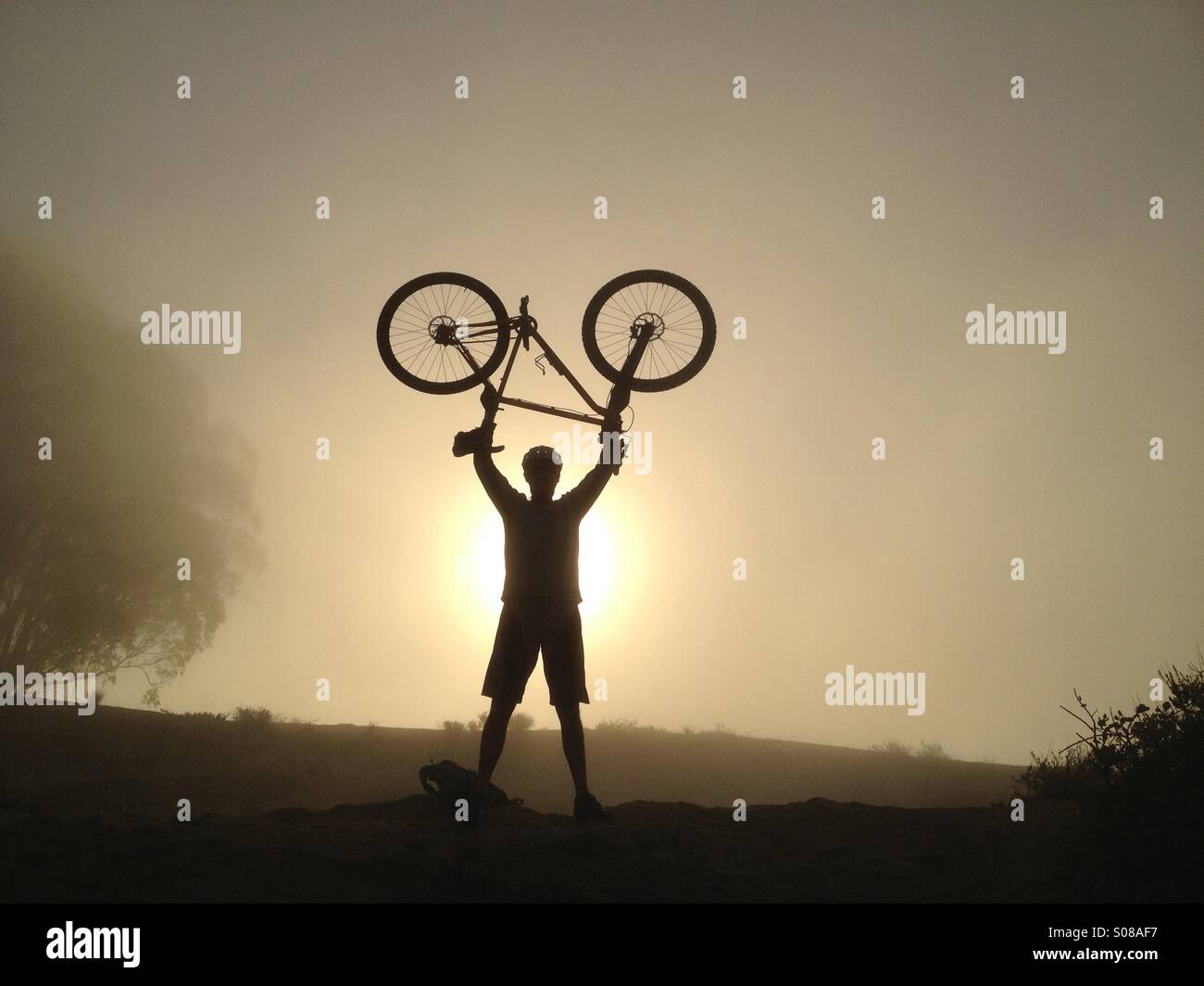 A mountain biker holds his bike over his head in silhouette at sunrise on a foggy morning in Laguna Beach, California, USA. Stock Photo