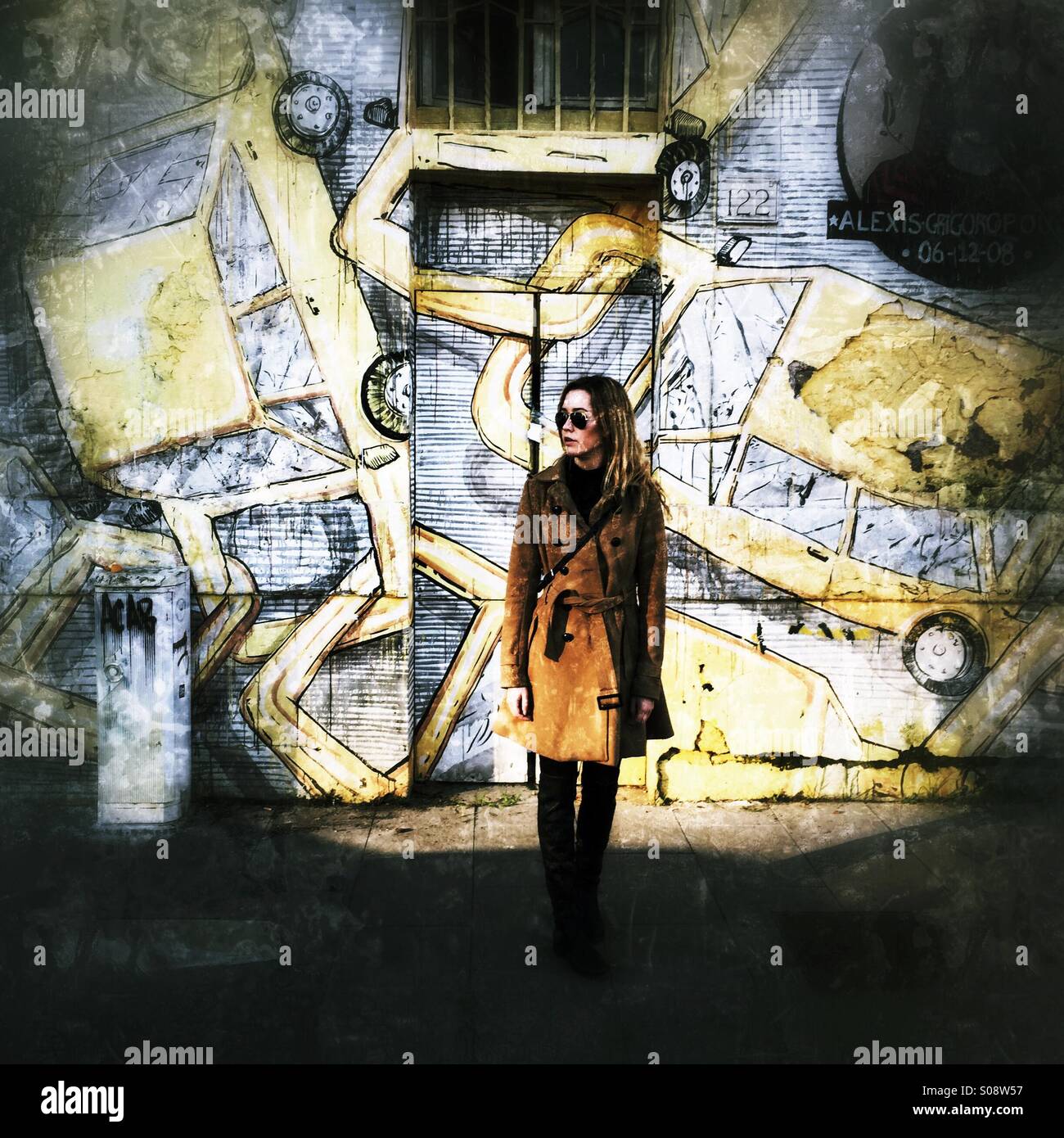 Modern high contrast image of young woman in a long brown coat in front of a wall with paintings of yellow cars on it. - Smartphone Captured Stock Image