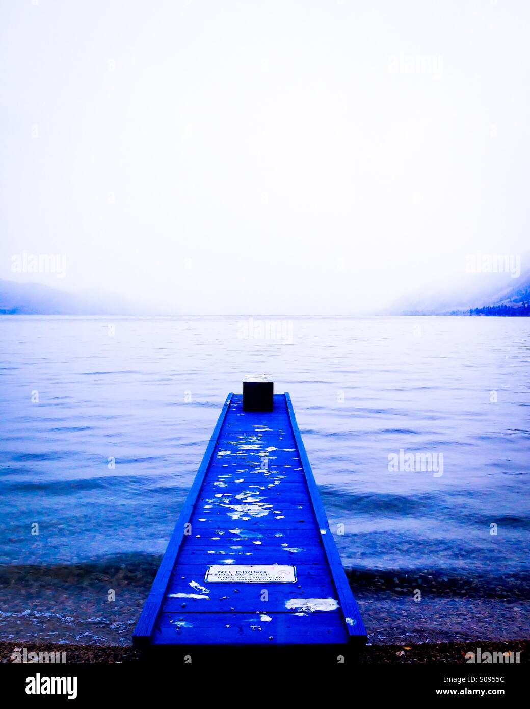 Blue fishing dock reaching into the cold waters of lake on early winter day. Mountains in the distance are engulfed in low clouds. - Smartphone Captured Stock Image