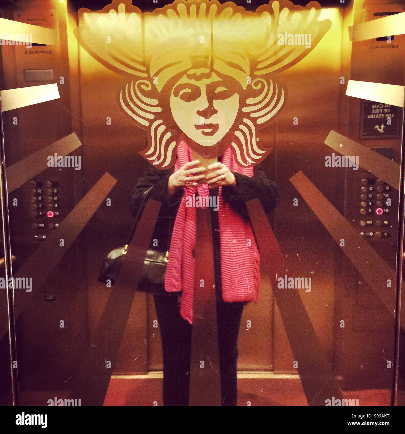A woman takes a comical selfie in an elevator that has the ornate head of a woman on the elevator wall. - Smartphone Captured Stock Image