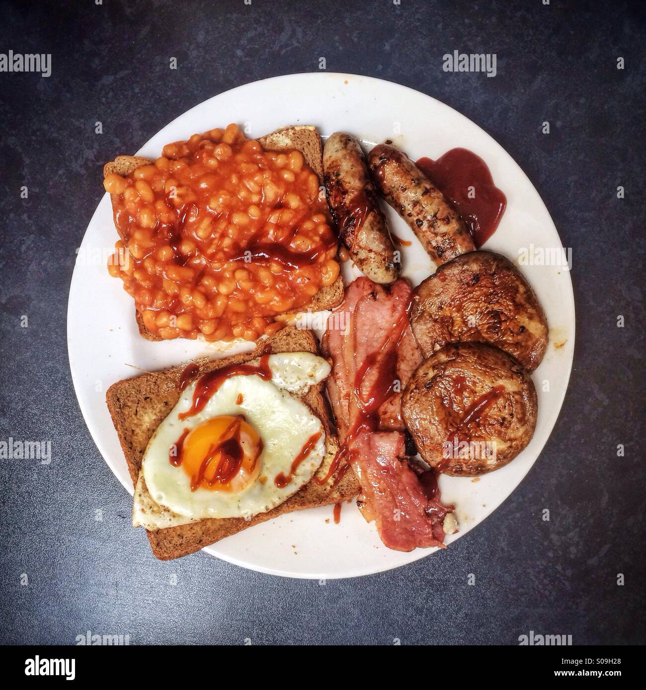 A full English breakfast of bacon, fried eggs, sausages, baked beans, toast and mushrooms with brown sauce on a white plate against a black background. Stock Photo