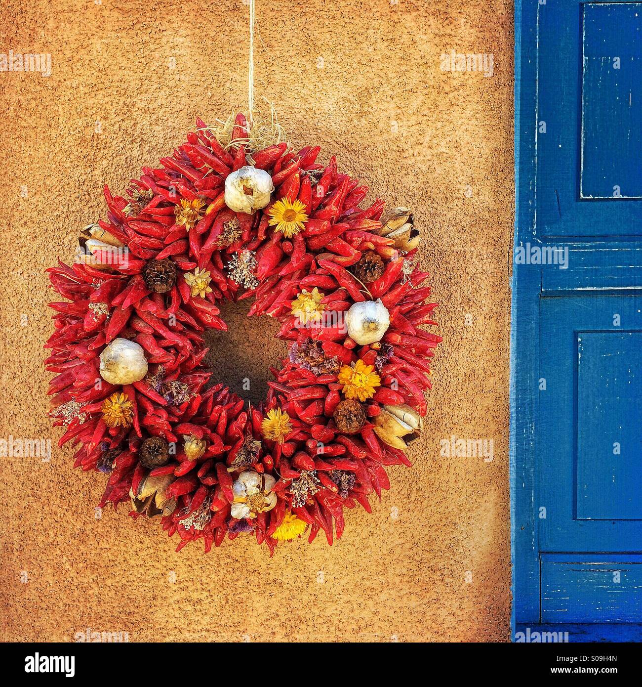 Red chile wreath hangs on stucco wall in the American Southwest next to blue door. Stock Photo