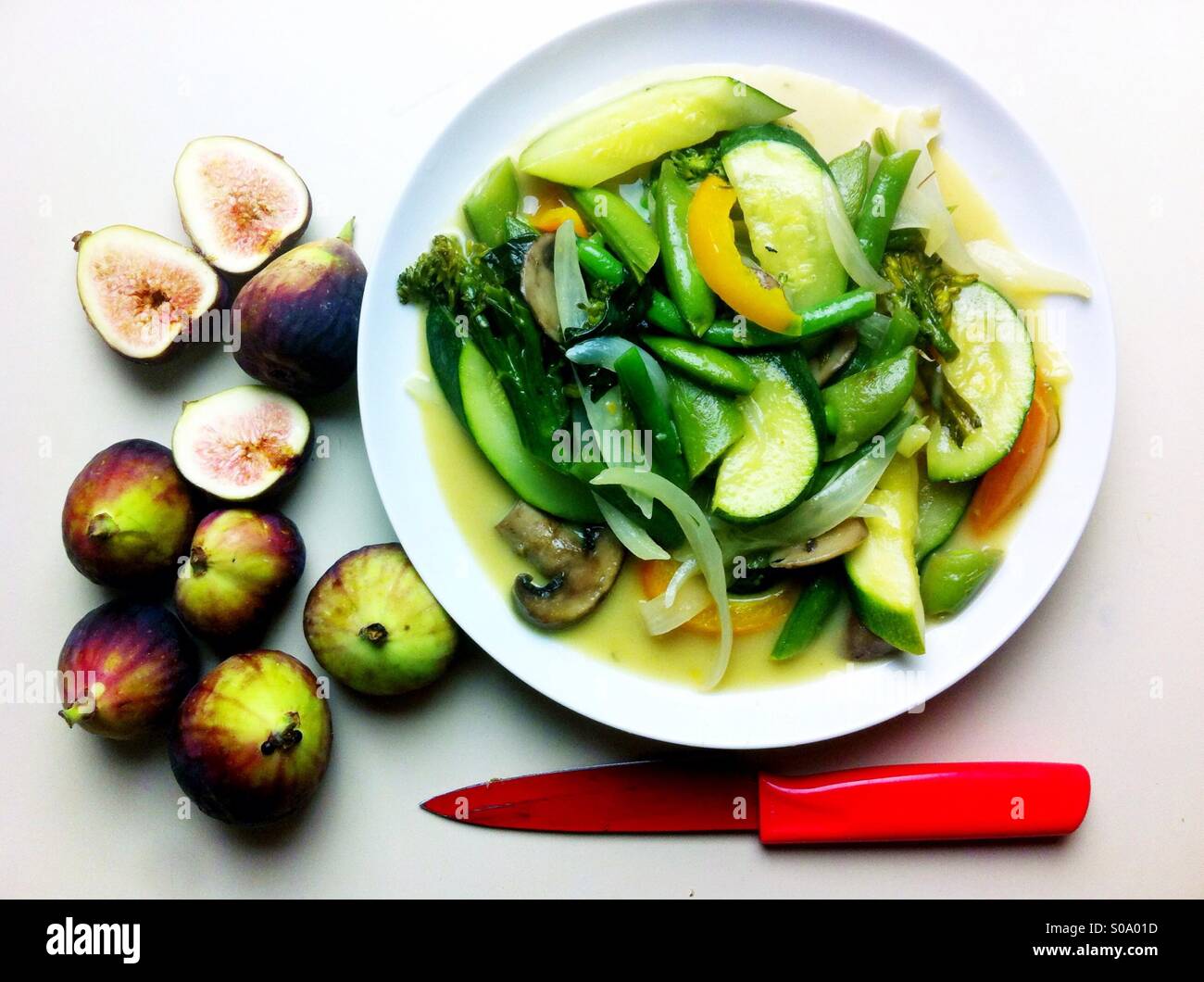 Vegan meal on white round plate with zucchini, bell peppers, onion, snow peas and fresh figs accompanied by the red paring knife Stock Photo