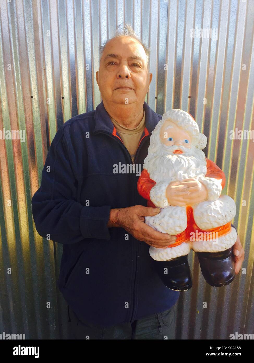 A 83-year-old man holding a plastic Santa Claus in front of a metal corrugated fence. Stock Photo