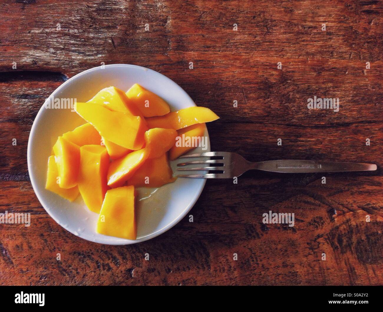 Pieces of yellow mango flesh on a white dish on distressed dark red wood table with metal fork Stock Photo