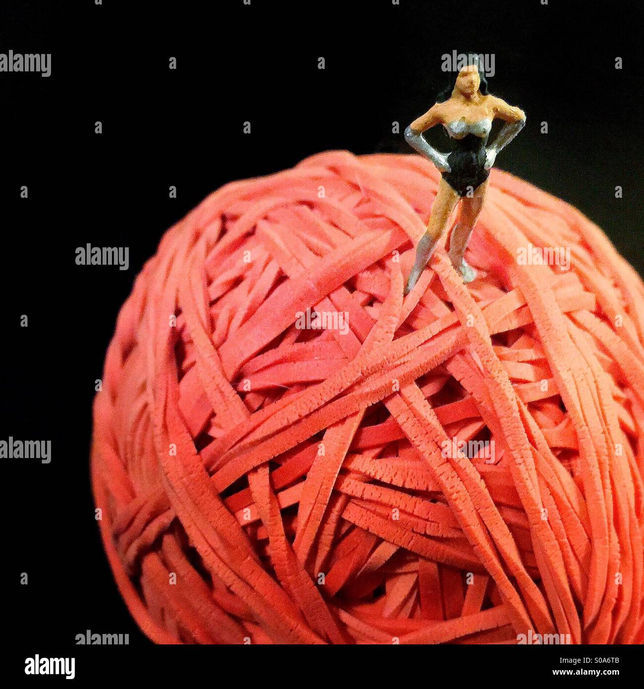 A small figure of a woman dressed like a superhero on top of a large ball made of red rubber bands - Smartphone Captured Stock Image