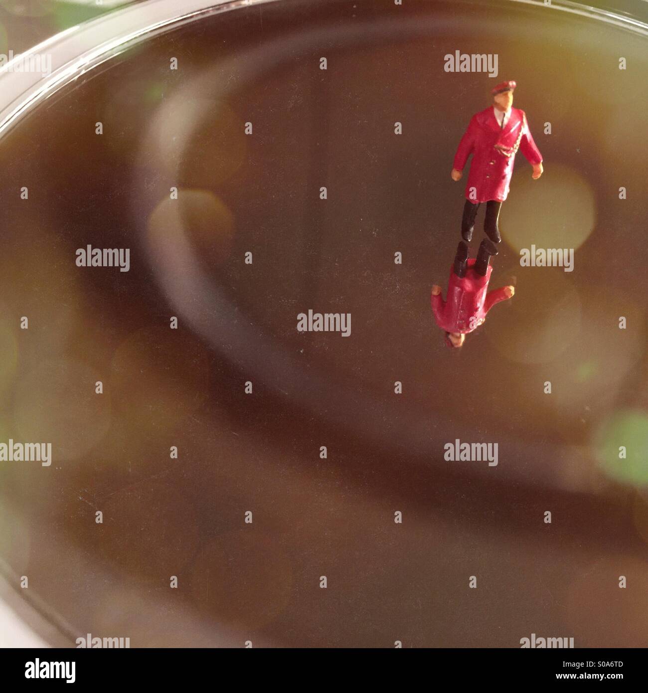 A tiny figure of a man in a red coat and a hat stands on a mirror above his reflection - Smartphone Captured Stock Image