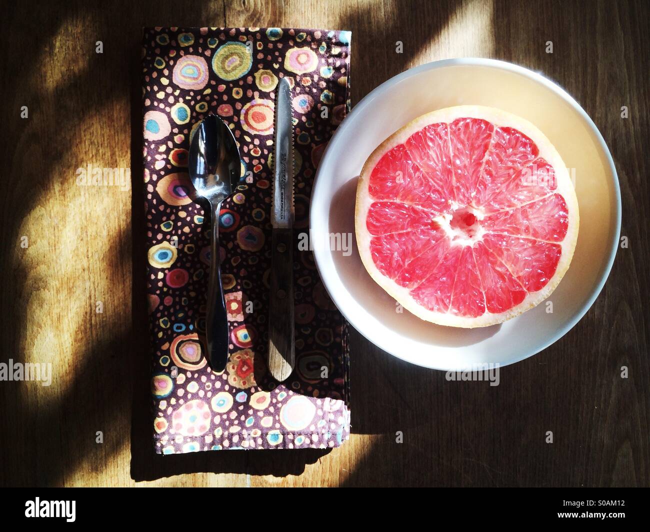 A red grapefruit, cut in half in a bowl with a cloth napkin, grapefruit spoon and a grapefruit knife on a wooden table. - Smartphone Captured Stock Image