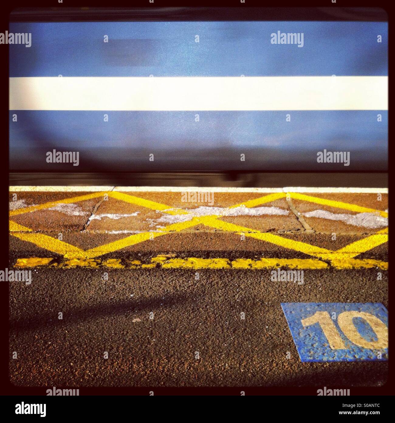 View of a station platform with a train passing through at high speed, yellow lines and the number ten painted in the foreground - Smartphone Captured Stock Image